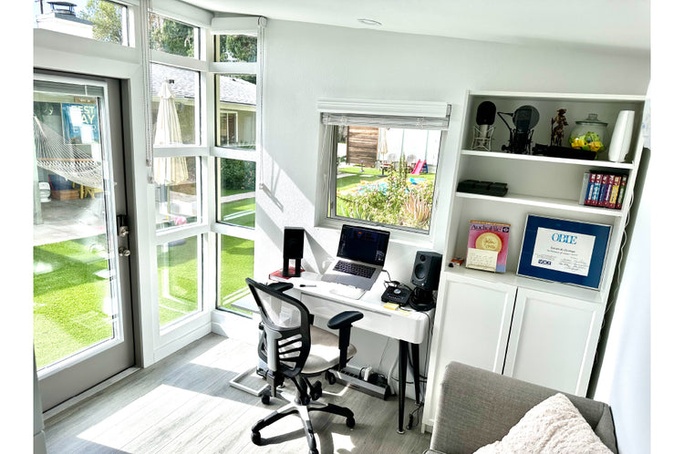 Home office with desk, chair, and bookshelf in a bright room with large windows.